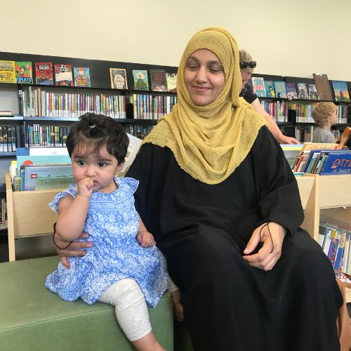 Mother with mustard coloured hijab sitting with daughter in blue patterned dress on green blocks, with children's books on shelves in the background