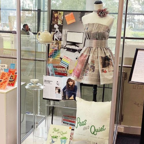 Large glass display case showing newspaper pleated dress on a mannequin, teapot and saucer, Peas and Quiet picture book and cushion cover