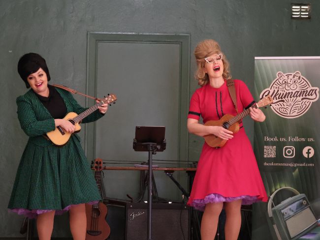 Dixie and Ronette Chandelle, both sporting impressive beehive hairdos, sing from the stage at Ladysmith, one a green checked dress smiling at the audience; the other in hot pink, eyes closed in song.