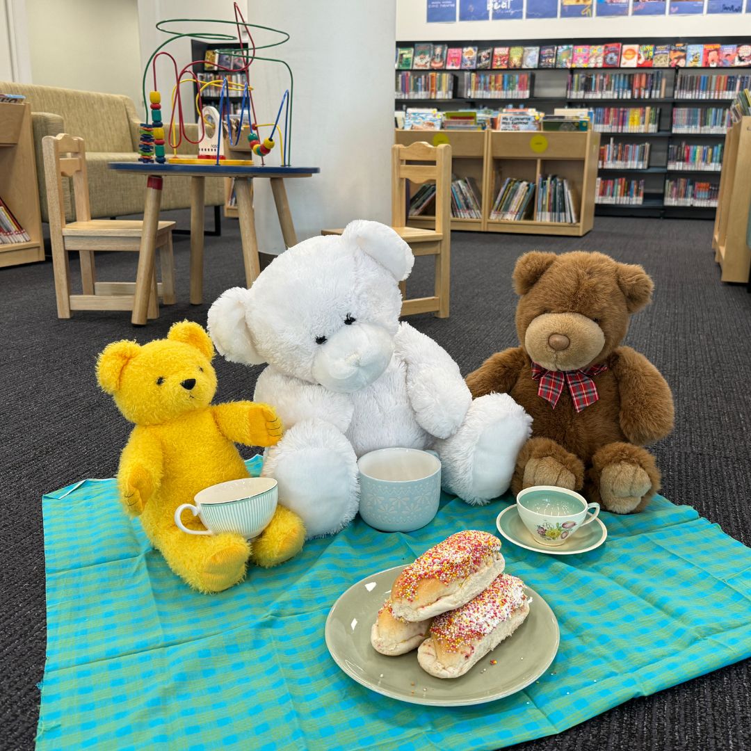 Three adorable teddy bears sit on a rug, with tea cups and finger buns on a plate