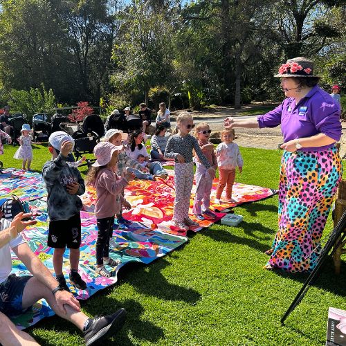 A librarian dressed in a purple top and colourful skirt leads a group of kids and grown-ups in the hokey-pokey.