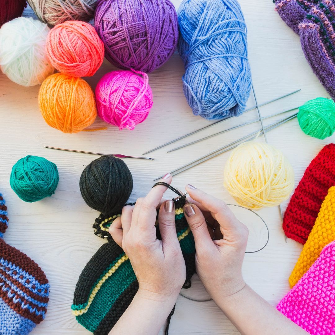 Colourful balls of wool are arranged around a table, with a pair of hands holding needles, knitting