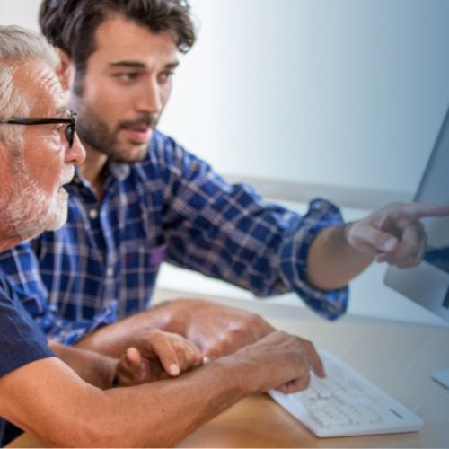 A younger bearded guy is helpfully showing an older bearded guy something on a computer monitor.