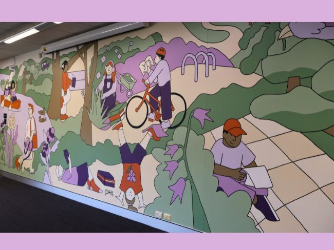 The mural, as seen from near one end, shows a park landscape featuring people reading while rowing in a river, sitting on a bike, gardening and playing.