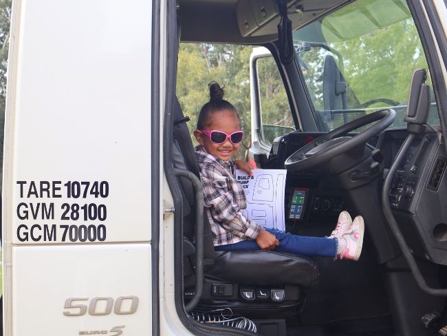 An exceptionally cool-looking kid in bright pink sunglasses is beaming from the driver's seat in the cab of a truck.