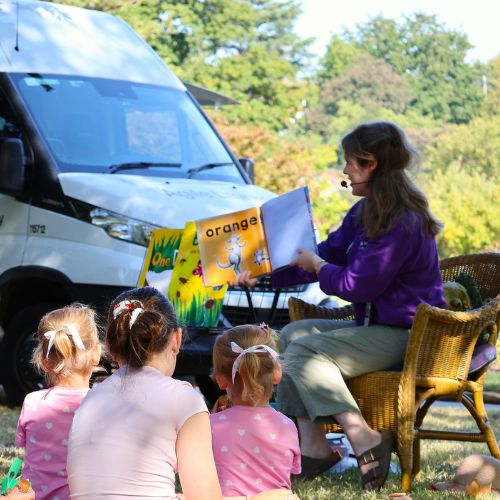 Children are seated before a grown-up storyteller, with the Agile van in the background
