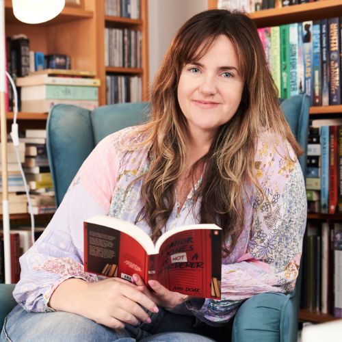 Amy has long brown hair, a cool floral top, and an excellent array of books in shelves behind her