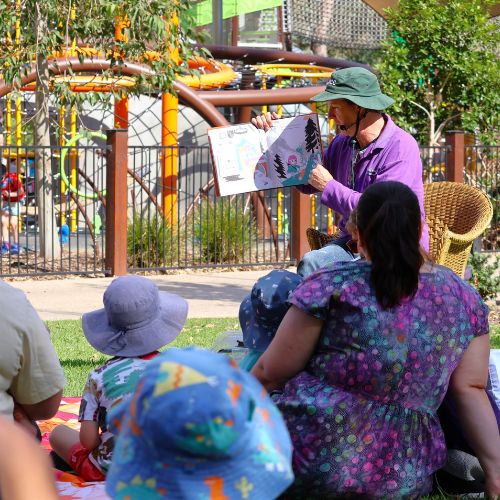 With an adventure playground in the background, storytime kids and their grownups listen to Peter reading a picture book