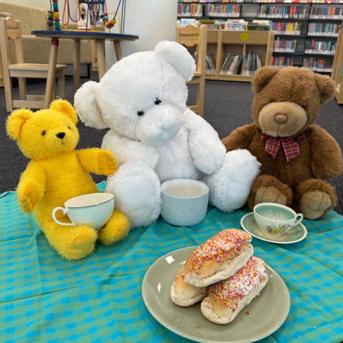 Three adorable teddy bears sit on a picnic blanket with teacups and fingers buns