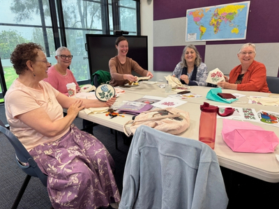 A group of women in the library gathered around a table for Stitch Social, talking and showing their craftwork.