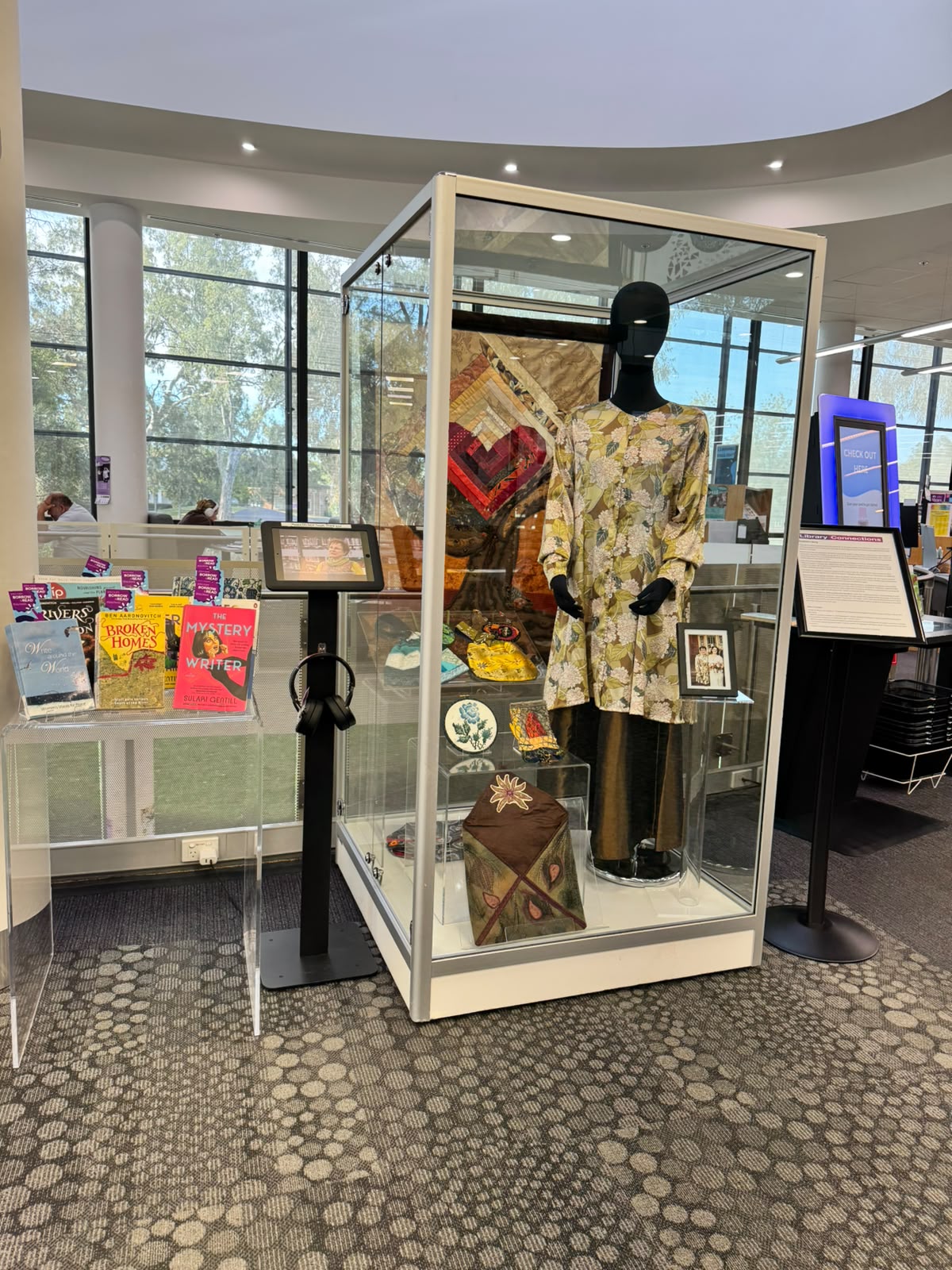 A photo of the Library Connections exhibition featuring Elizabeth's story. This includes a shelf displaying her favourite books, and a large glass case showcasing her handmade craft pieces.